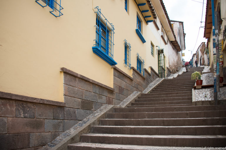 Long steep stone staircase rising between yellow and white buildings with blue window trim.