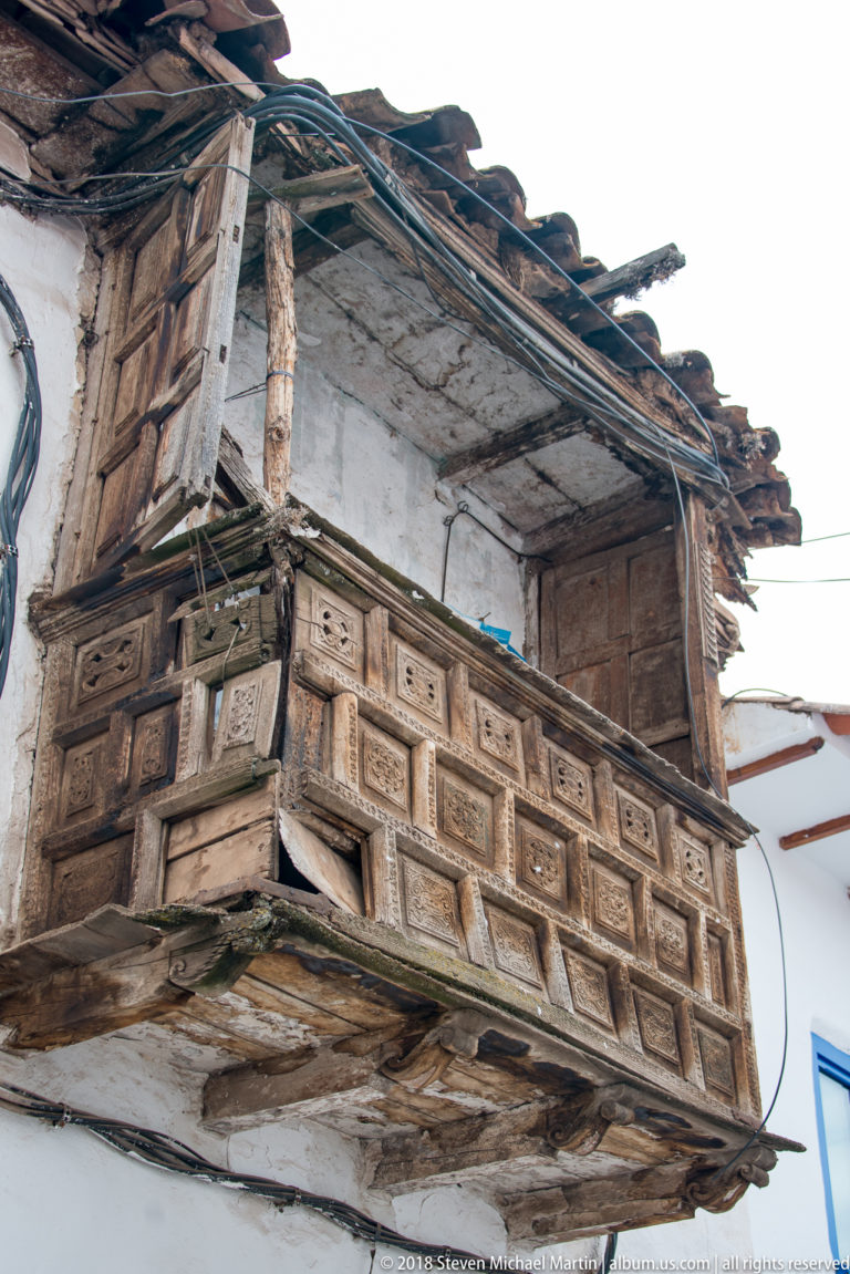 Weathered projecting wooden balcony with carved panels beneath clay roof tiles on a white wall.