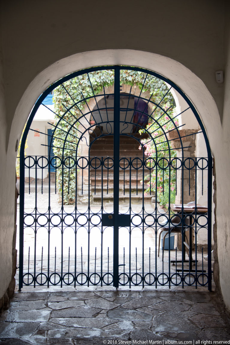 Black wrought-iron gate beneath a rounded arch, looking into a planted courtyard.
