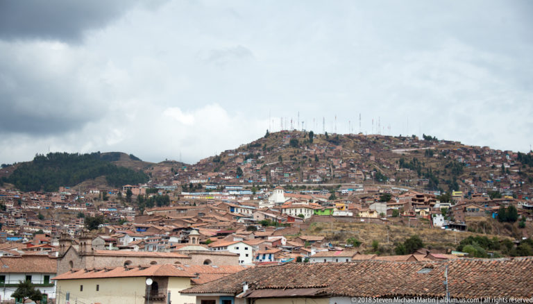 Wide Cusco roofscape beneath dark clouds, with dense hillside neighborhoods and communications towers beyond.