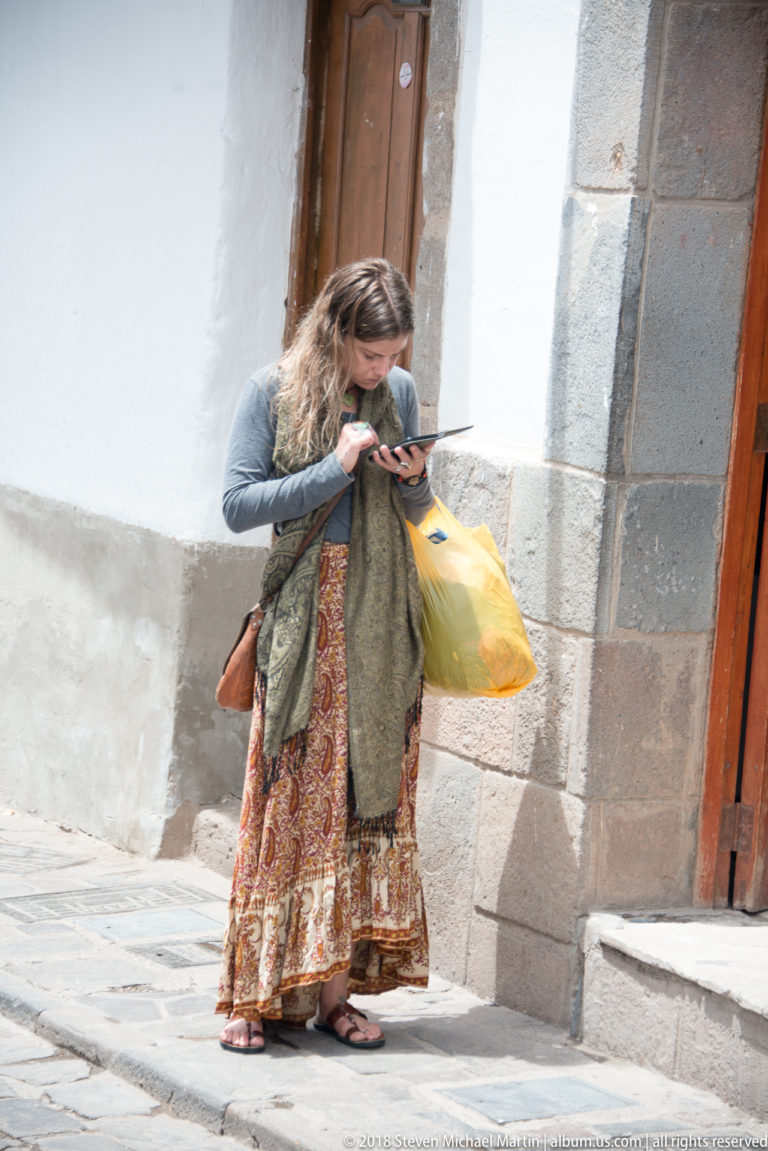 Woman in patterned skirt standing beside a stone doorway on a sunlit Cusco street.