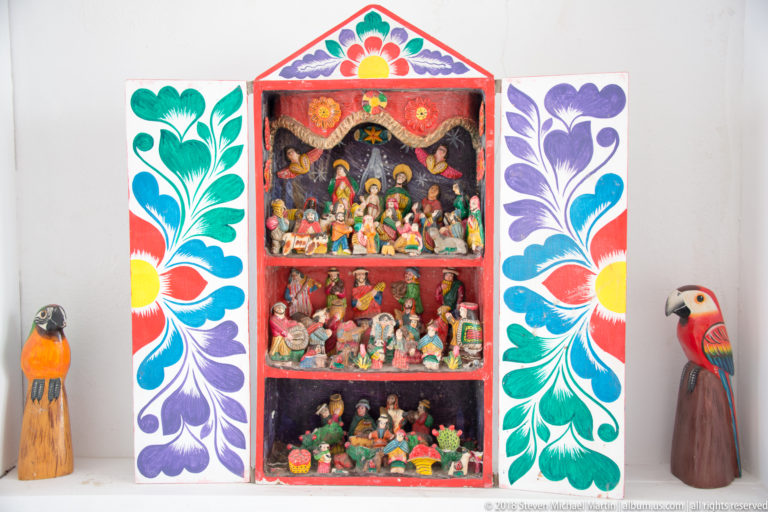 Open painted retablo with three tiers of miniature figures, floral doors, and carved bird figures beside it.