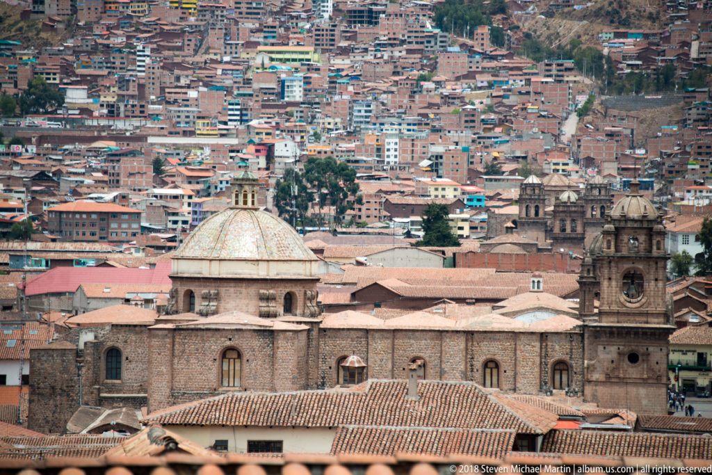Large stone church with pale dome and bell tower against densely built Cusco hillsides.