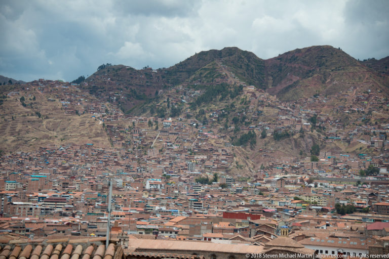 Wide Cusco cityscape of dense hillside neighborhoods climbing toward rugged mountains beneath cloudy sky.