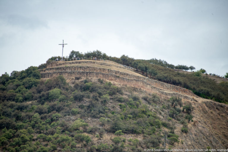 Curving stone terraces on a scrubby hillside below a large cross at the summit.
