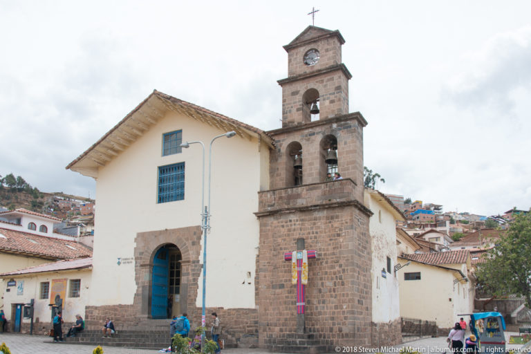 Whitewashed church with blue door and a tall stone bell tower topped by a clock and cross.