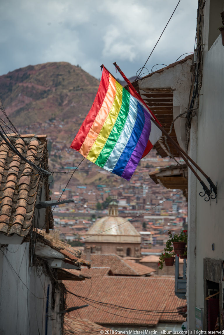 Rainbow-striped flag above tiled roofs with densely built Cusco hillside in the background.