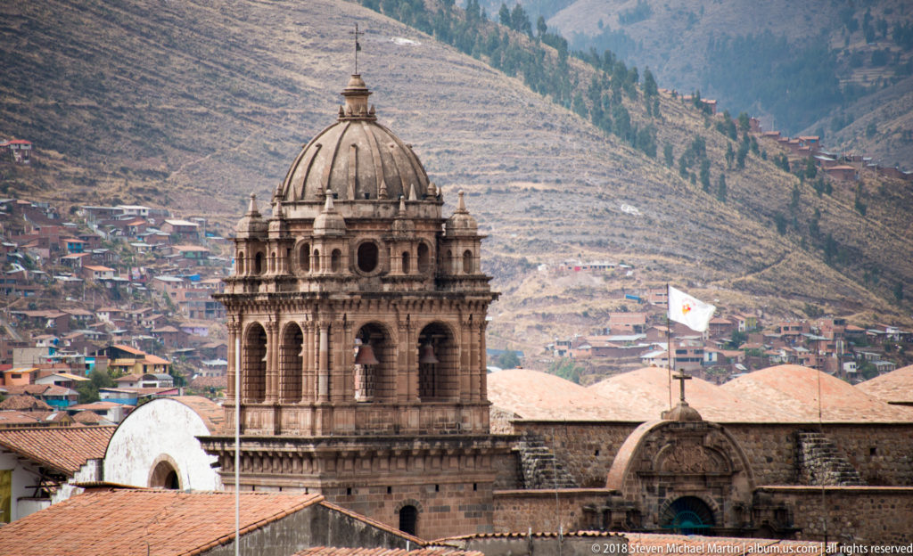 Ornate stone bell tower with arched openings and ribbed domed cap against a densely built hillside.