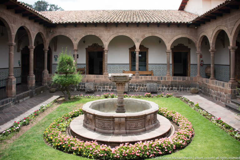 Arcaded colonial courtyard with a circular stone fountain, lawn, flowers, and tiled roof.