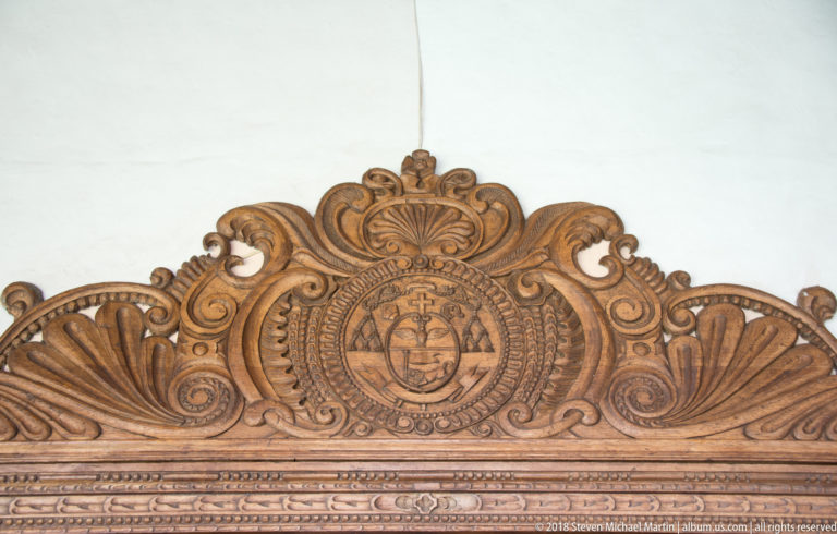 Ornate carved wooden crest with scrolls and a central shield mounted above a doorway.