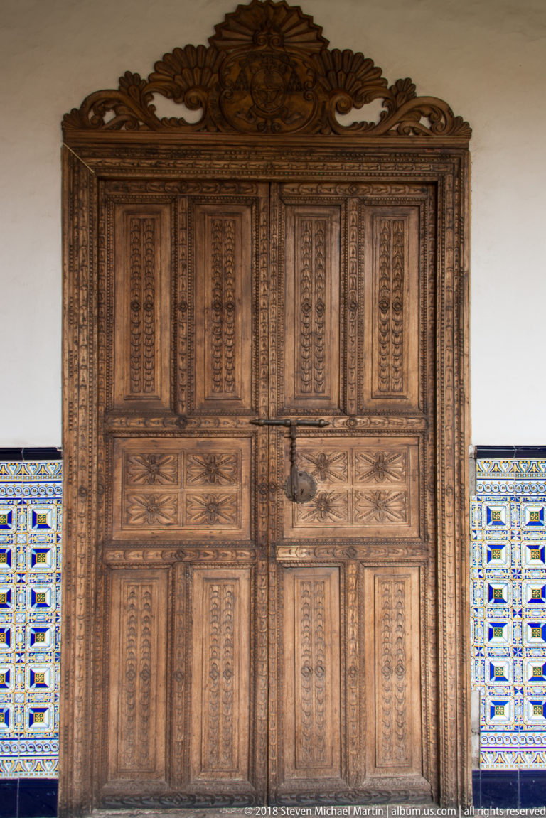 Tall carved wooden double doors with paneled relief and an ornamental crest, flanked by patterned tilework.