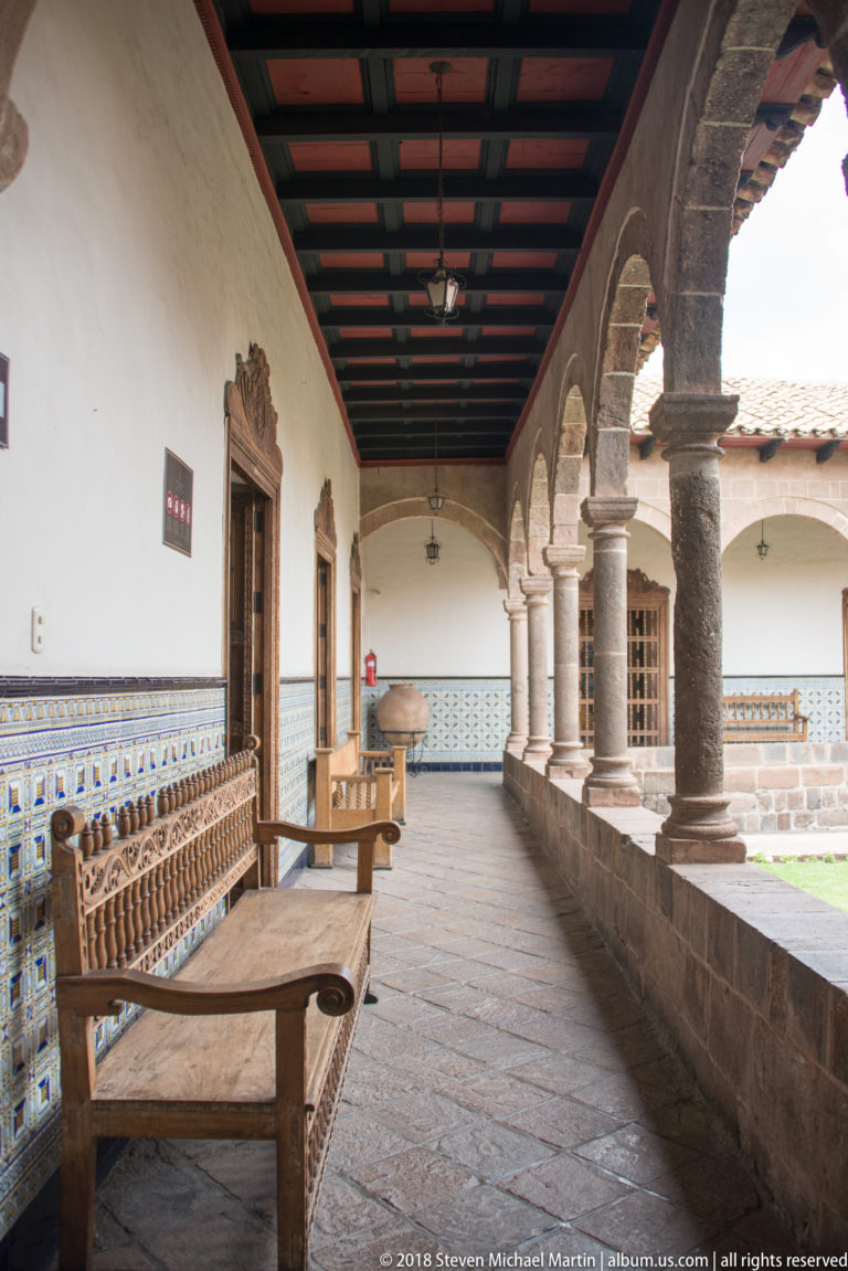 Covered historic corridor with stone arcade, patterned blue-and-white tile wainscot, timber ceiling, and wooden bench.