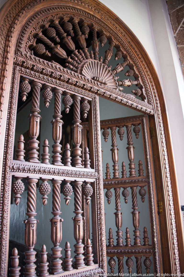 Ornate arched wooden door or screen with turned balusters, spiral elements, and dense carved floral decoration.