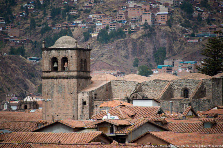 Massive square stone bell tower beside a long church, with tiled roofs and hillside housing behind.