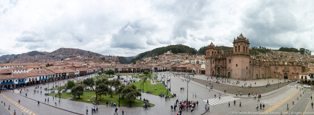 Elevated panorama of Cusco’s Plaza de Armas with lawns, cathedral, arcades, and steep hillside neighborhoods.
