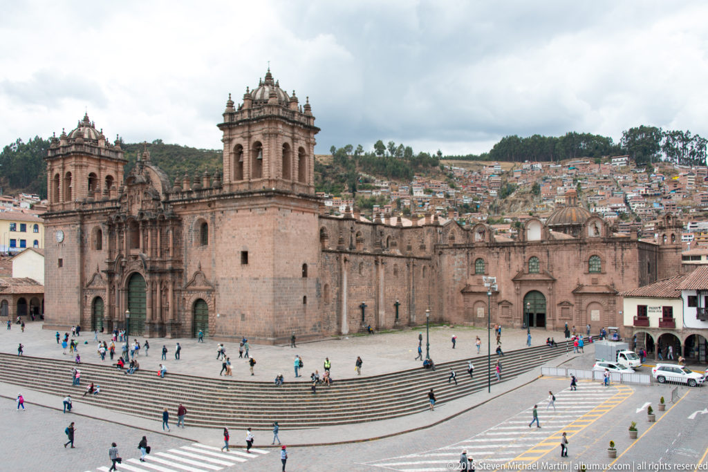 Wide view of Cusco Cathedral beside the Plaza de Armas, with pedestrians and hillside neighborhoods beyond.