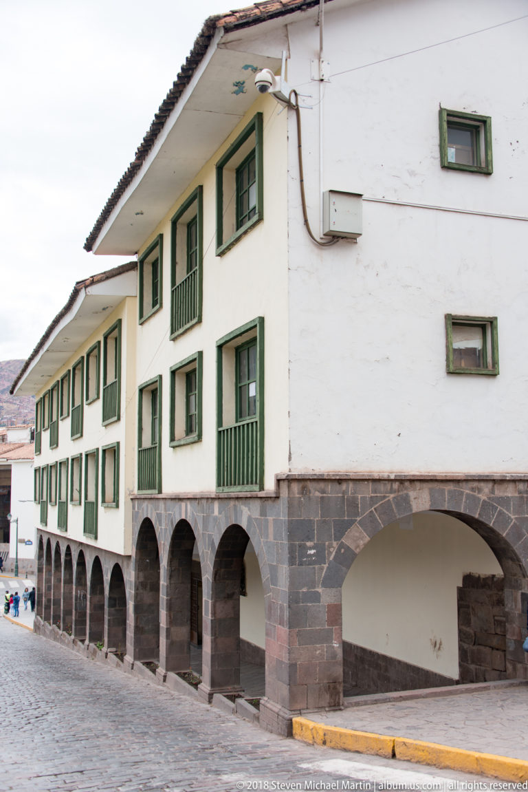 White historic building with green windows and balconies above a dark stone ground-floor arcade.