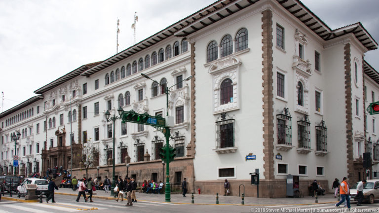 Long white historic building with stone trim, arched upper windows, balconies, pedestrians, and traffic.