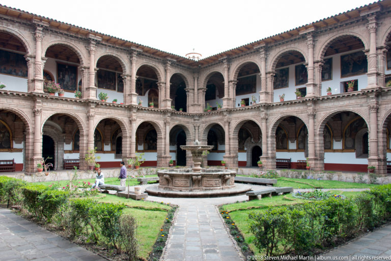 Corner view across a landscaped two-story cloister courtyard toward a central stone fountain.