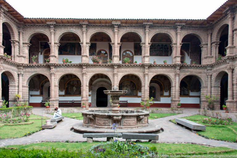 Two-story arcaded courtyard with a central stone fountain, lawns, flower beds, and surrounding galleries.