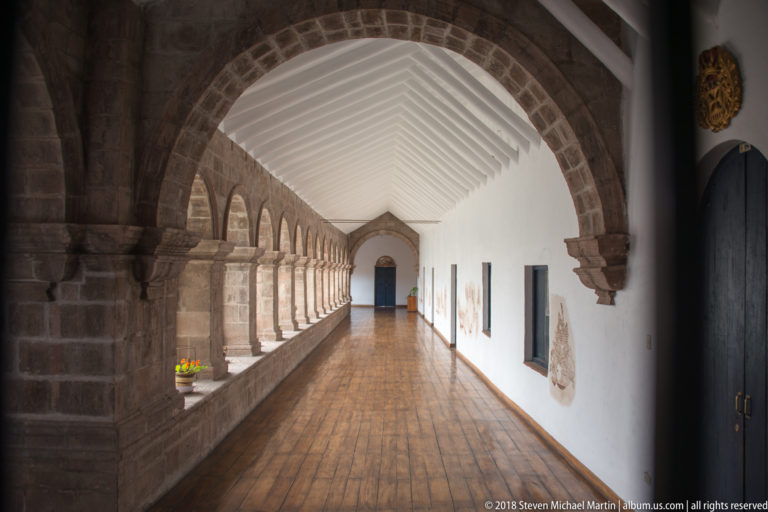 Long arcaded gallery with stone arches, white timber ceiling, wooden floor, and a doorway at the far end.