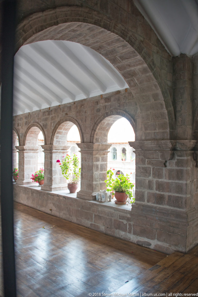 Stone cloister corridor with broad arched openings, white roof beams, and potted flowers.