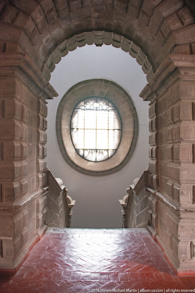 Stone steps pass beneath masonry arches toward a large oval window in a white interior wall.