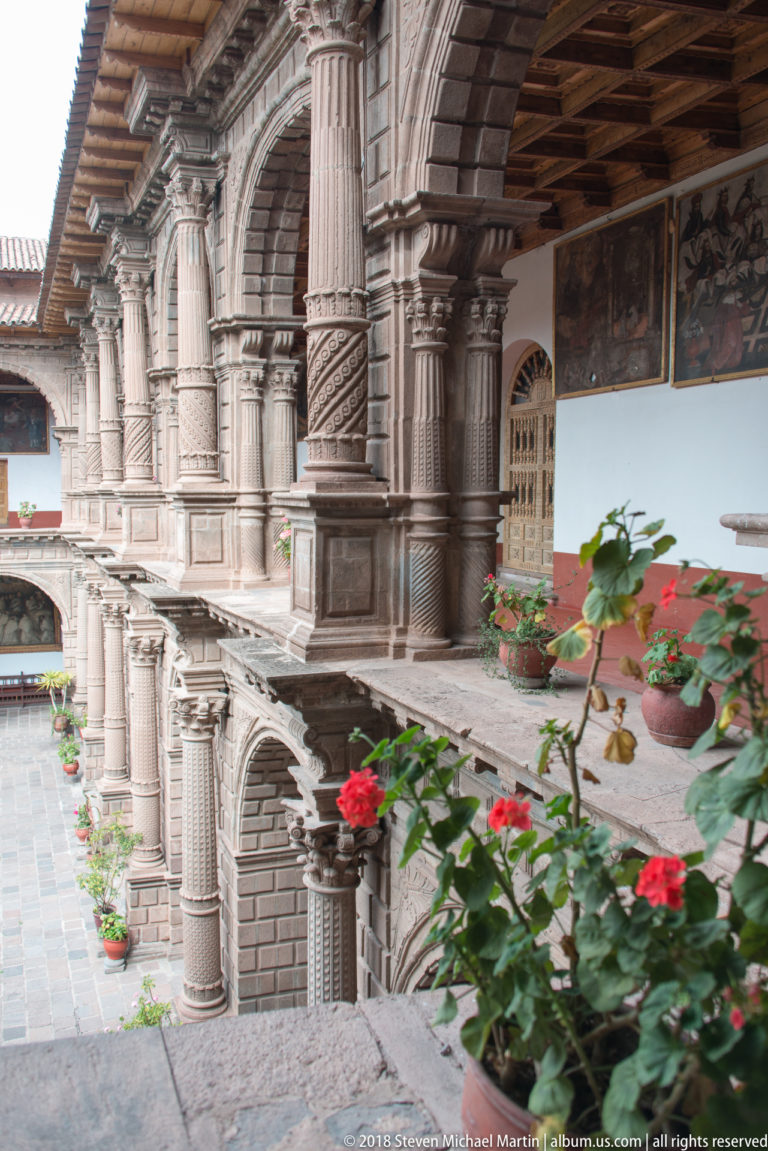 Upper cloister walkway lined with carved stone columns, arches, and flowering potted plants.
