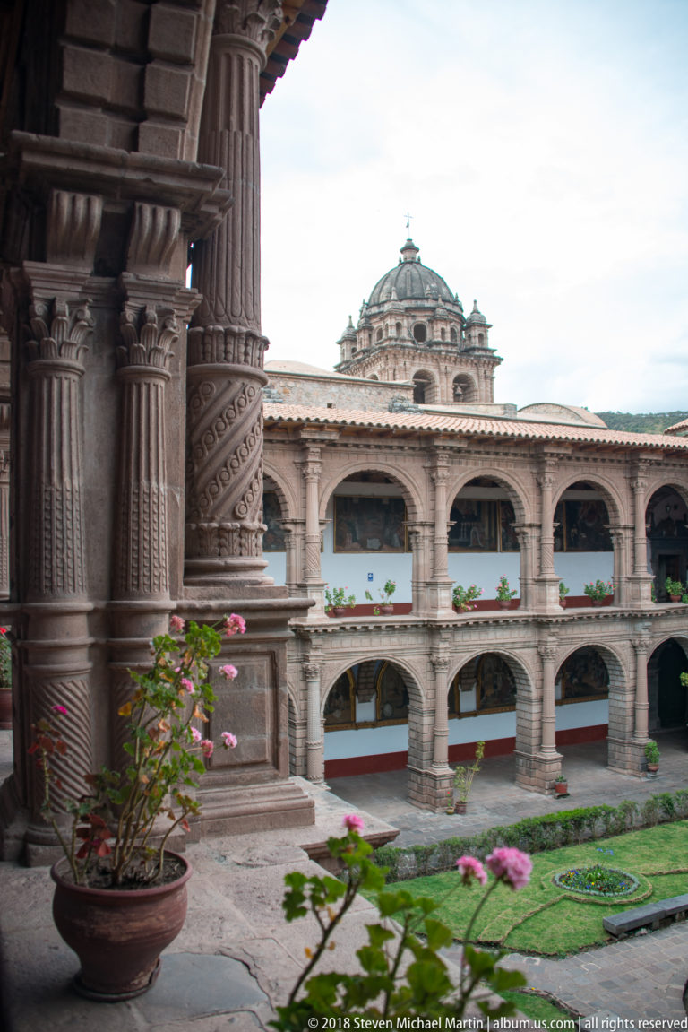 Two-story stone cloister with garden beds, carved columns, and a domed church rising beyond.