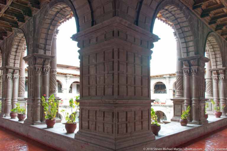 Large carved stone pier and arches open onto a bright cloister courtyard with potted plants.