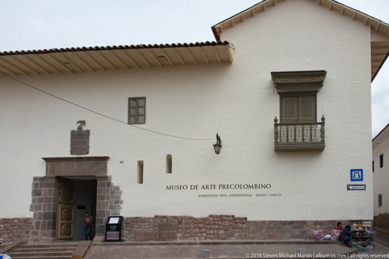 White exterior façade of Museo de Arte Precolombino with stone base, carved entrance, projecting balcony, wall sign, and pedestrians at street level.