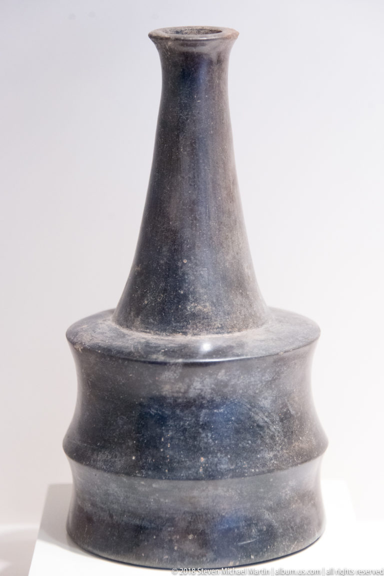 Dark burnished ceramic vessel with tall flared neck and three projecting stepped bands around its broad lower body.