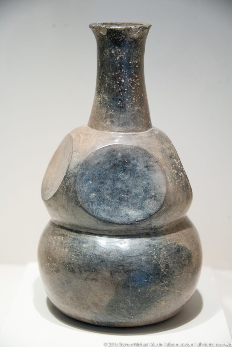 Dark ceramic pitcher with narrow flaring neck, stacked rounded bands, projecting shoulder, and a large smooth oval panel on the body.