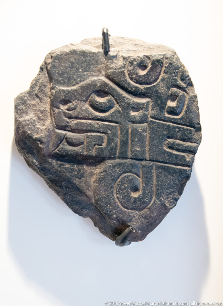 Fragmentary dark stone slab with deeply incised symmetrical face-like design, large eyes, broad mouth, and curling fanged elements.