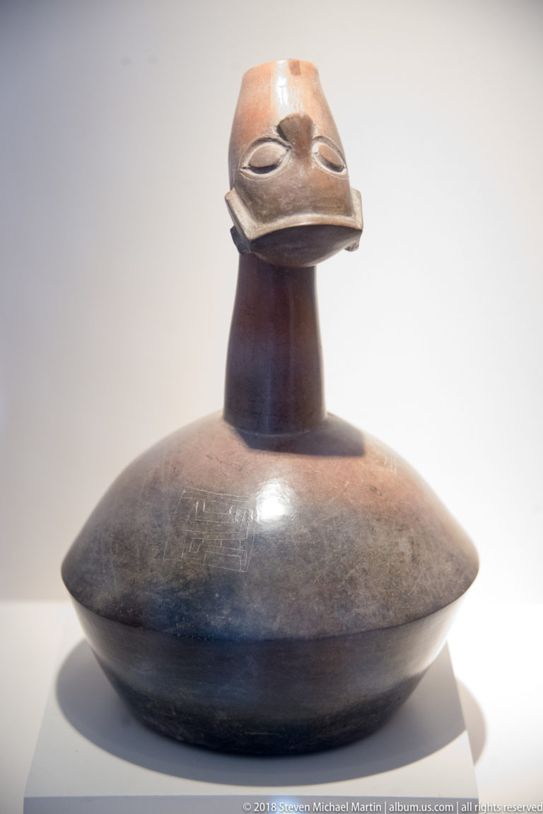 Dark burnished ceramic bottle with flattened round body, tall neck, and a modeled human head at the mouth.