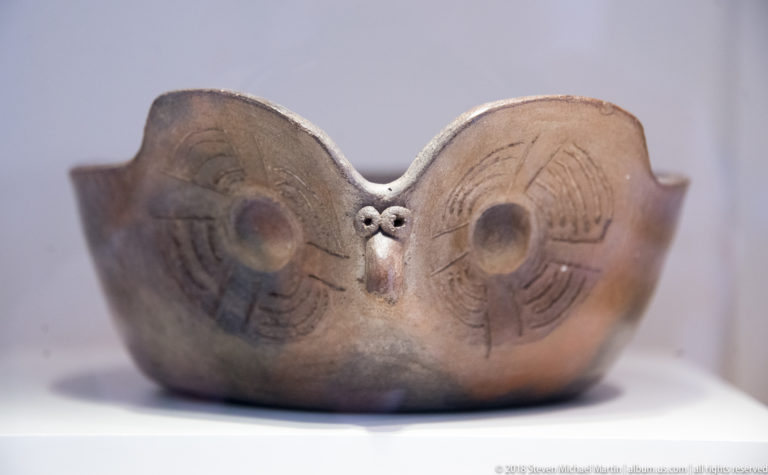 Shallow dark ceramic bowl modeled as an owl face, with large circular eyes, incised arcs, small beak, and raised side lobes.