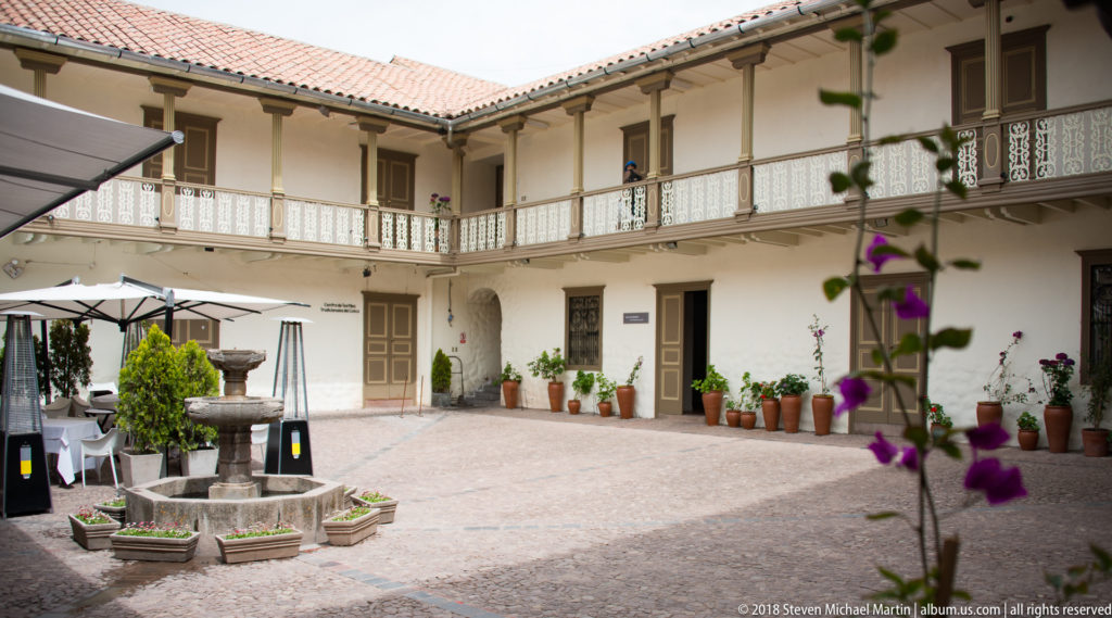 Wide museum courtyard with two-story balcony, white walls, potted plants, central stone fountain, planters, and outdoor seating.