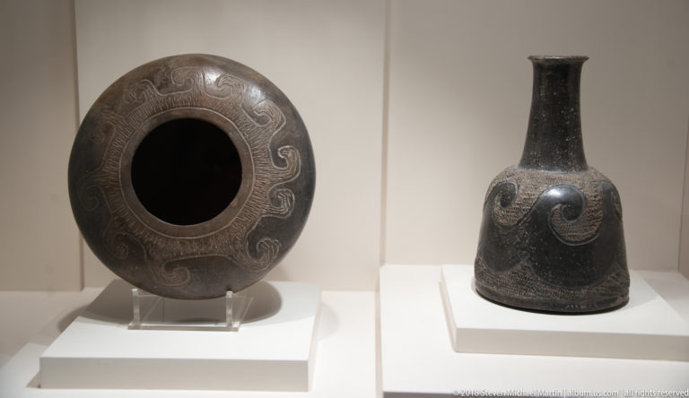 Dark ceramic ring vessel with incised curves beside a tall narrow-necked bottle bearing a large spiral-like motif.