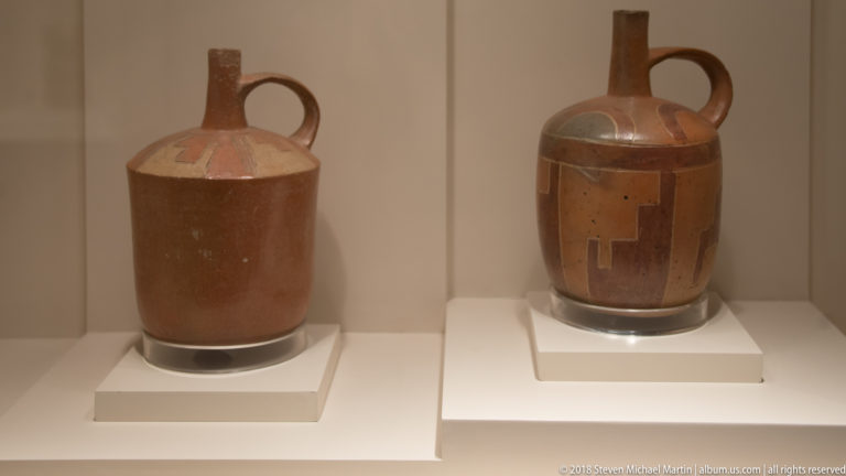 Two brown ceramic vessels on white plinths, one shorter with loop handle and one taller with long spout and painted blocks.