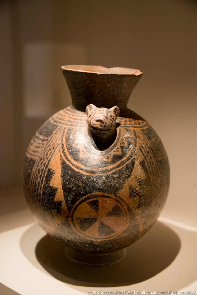 Low globular ceramic bottle with black and red geometric painting, flaring neck, and small modeled animal-like head on the shoulder.