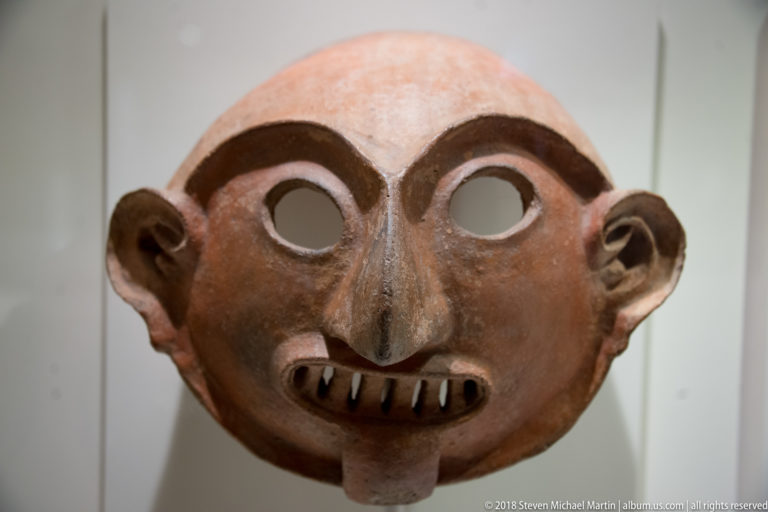 Reddish-brown ceramic mask with open eye sockets, heavy brows, narrow nose, projecting ears, and a tooth-filled mouth.