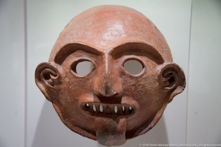 Reddish ceramic face mask with pierced eyes, arched brows, large ears, long nose, open mouth, and contrasting teeth.