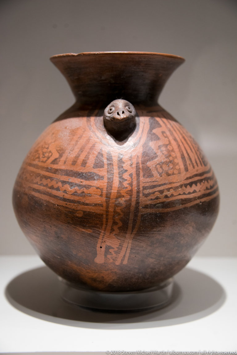 Globular painted ceramic bottle with broad dark neck, radial geometric bands, and a small modeled face on the shoulder.
