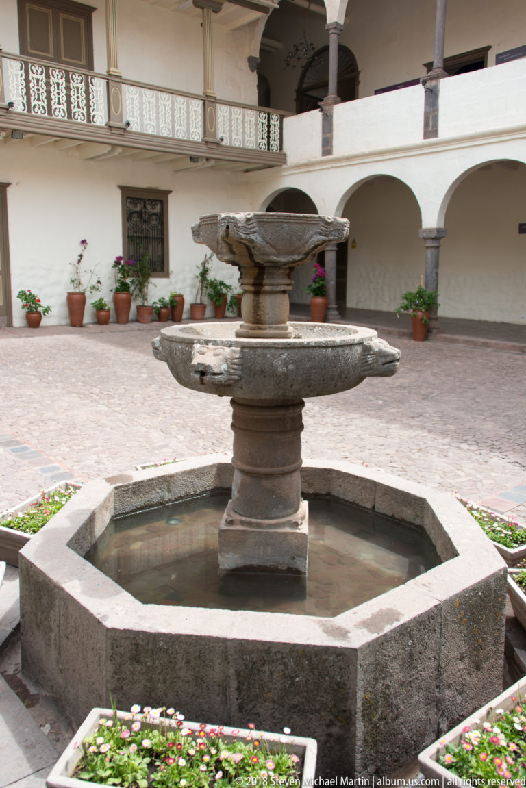 Stone fountain centered in an octagonal pool, surrounded by white arcades, stone columns, upper gallery, and potted plants.