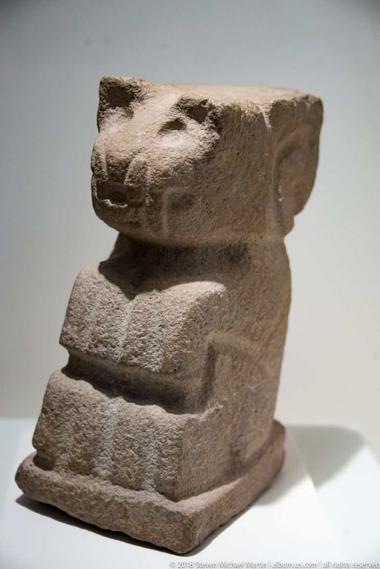Weathered crouching animal sculpture with projecting head, short muzzle, small eye, and blocky stepped body.