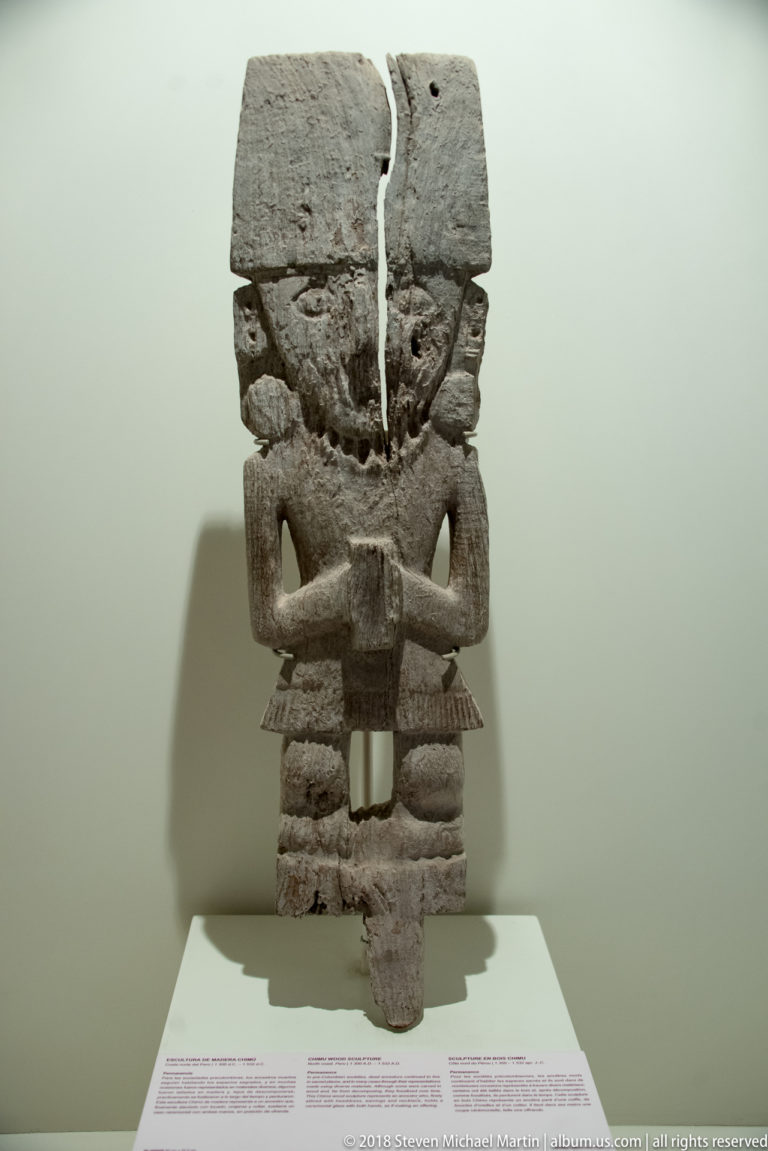 Front-facing wooden figure with a deep vertical head crack, large eyes, crossed arms, and blocky patterned legs.