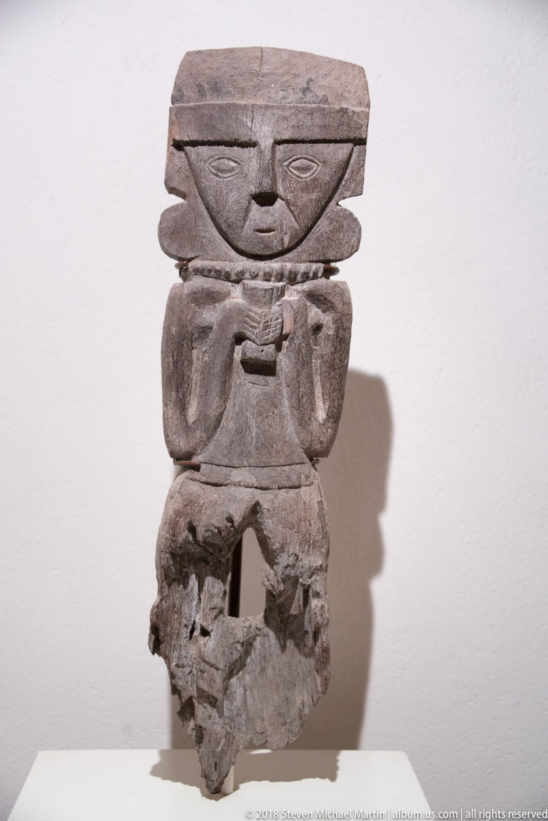 Wooden anthropomorphic figure with broad rectangular head, incised eyes, bent arms, and a small rectangular object at the chest.