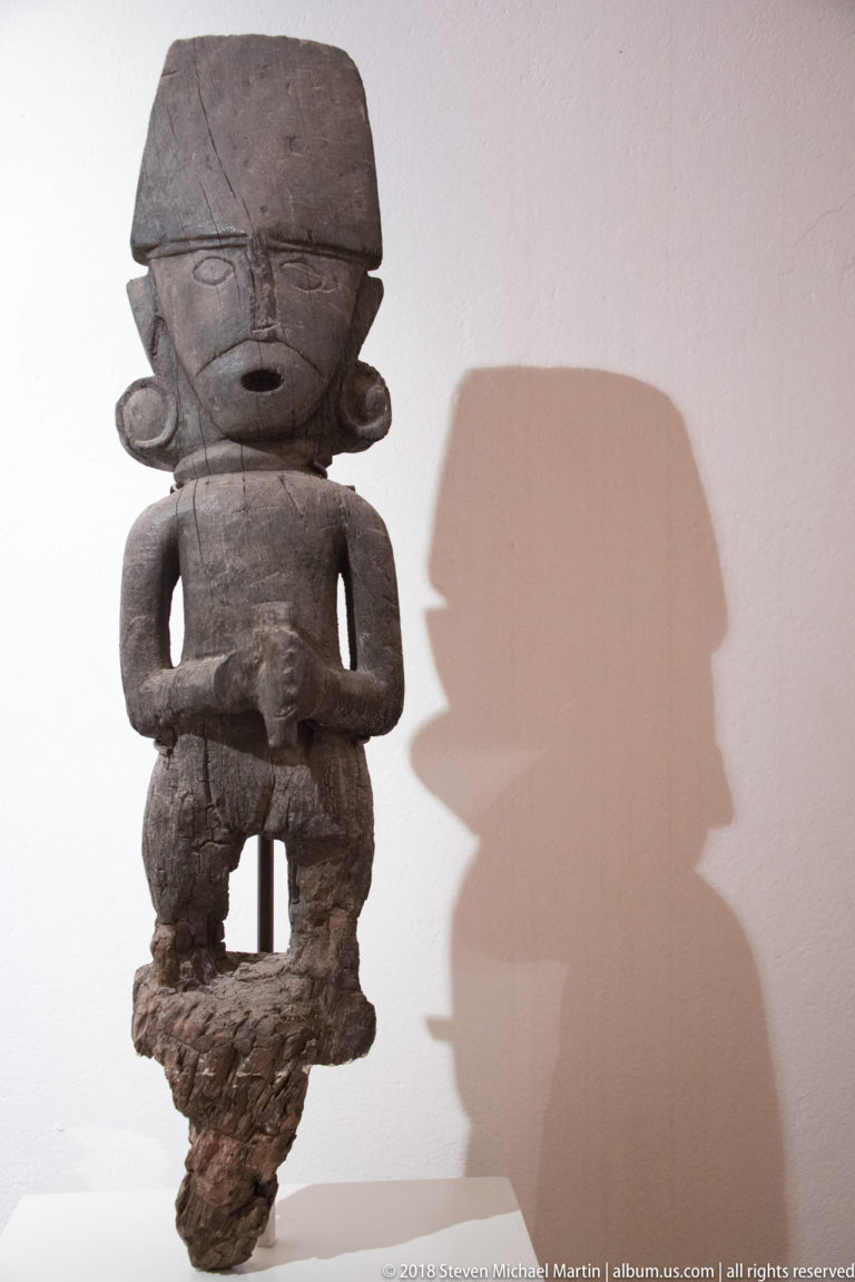 Dark wooden figure with oversized head, side projections, bent arms, short legs, rough lower extension, and large wall shadow.