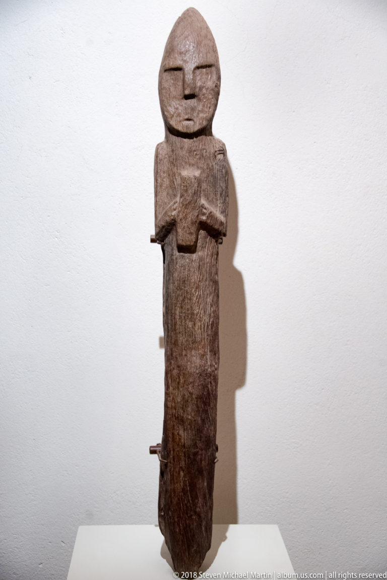 Tall narrow wooden standing figure with rounded head, slit eyes, folded arms, long body, and visible wood grain.