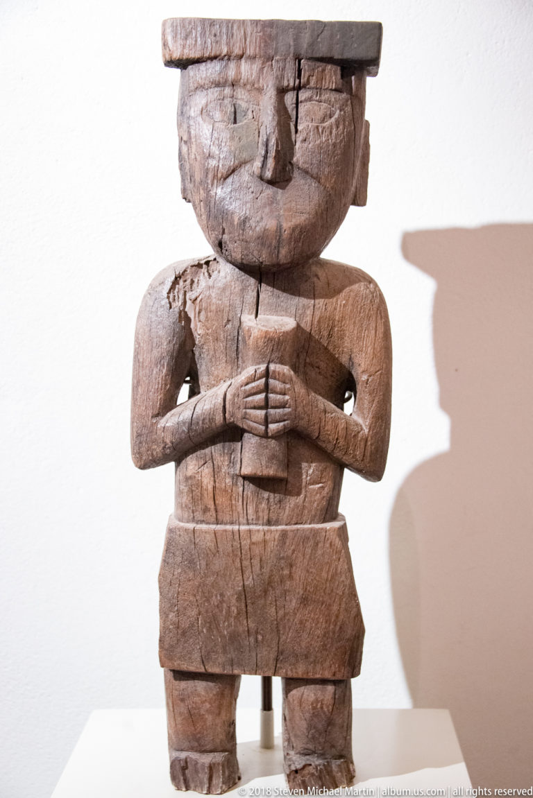 Large standing wooden figure with broad head, bent arms, hands clasped at the chest, short legs, and strong wall shadow.
